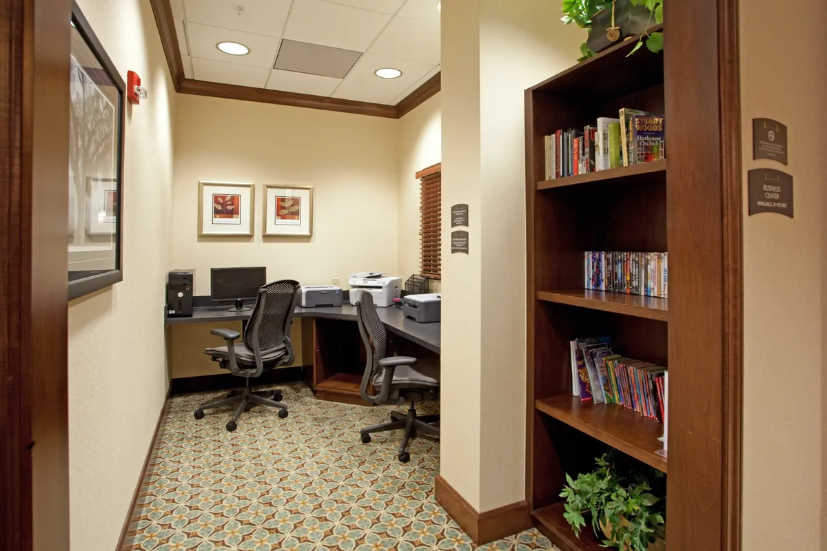 This image highlights a productive business center within the hotel, showcasing desks equipped with computers, a bookshelf stocked with books and DVDs, and framed artwork. The atmosphere is calm, allowing guests to work comfortably or engage in collaborative sessions.
