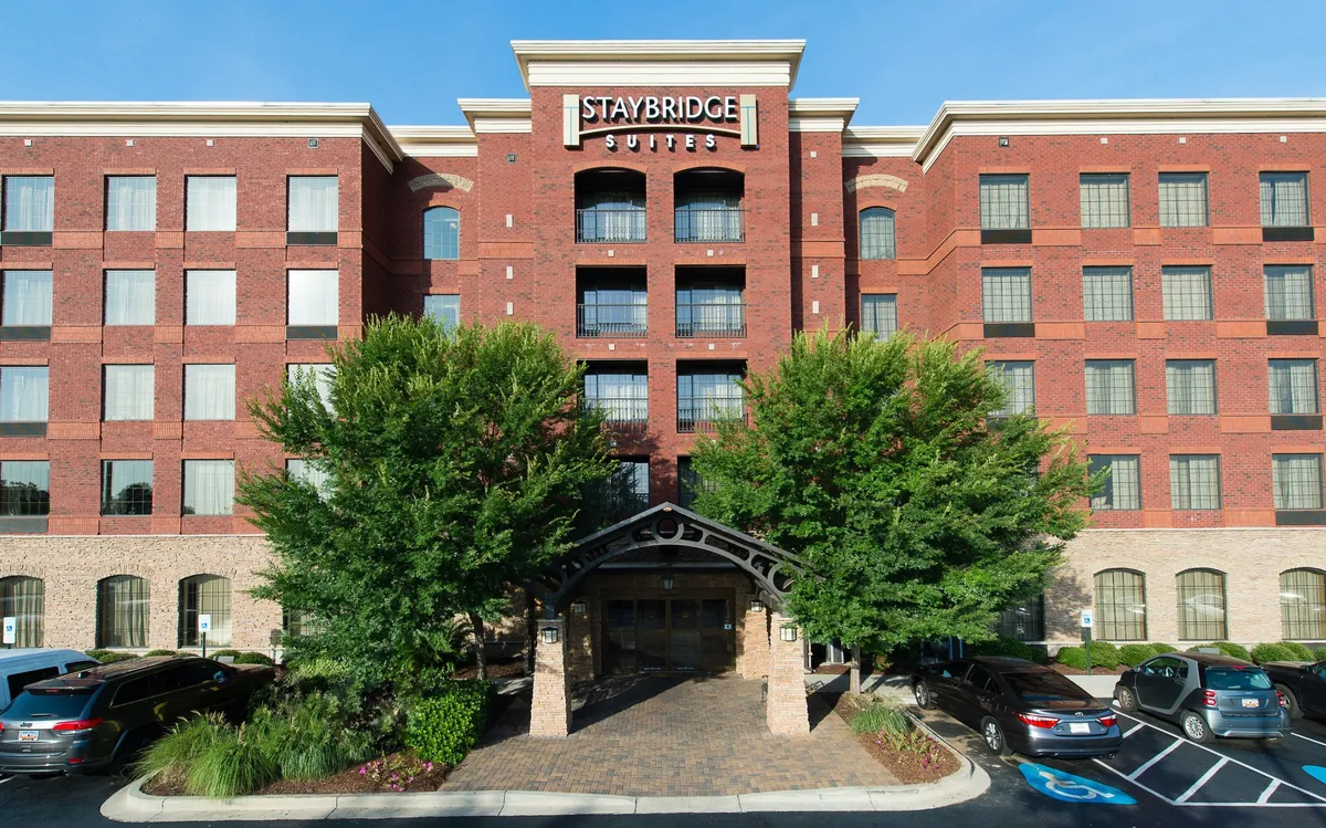 The image highlights the Staybridge Suites exterior, showing the welcoming entrance awning, prominent signage, and lush green trees framing the brick facade. The scene exudes approachability and hints at the hotel's proximity to downtown attractions, inviting guests to embark on their Columbia explorations right from the doorstep.