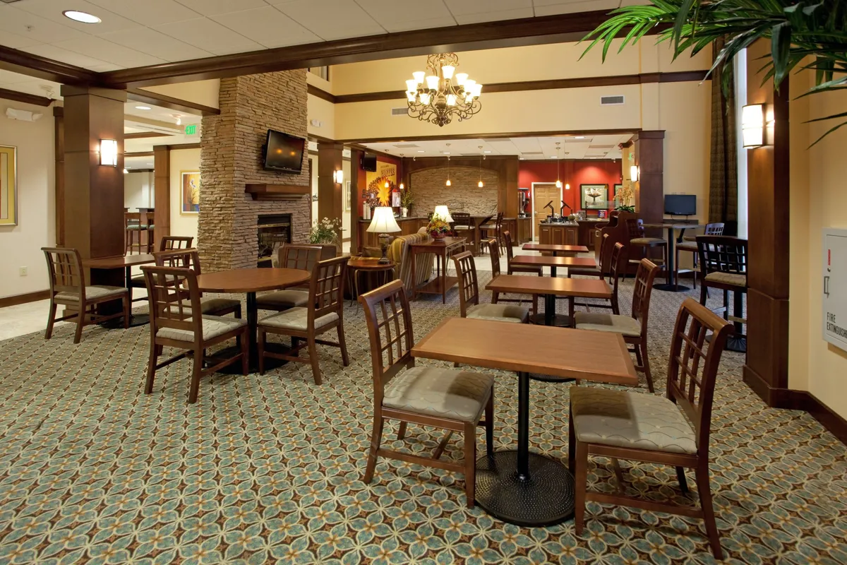 A spacious, warmly lit common area with wooden tables, a chandelier, decorative plants, and a fireplace. The seating is arranged to encourage both social gatherings and quiet moments, reflecting the suite's ability to adapt to different guest needs.