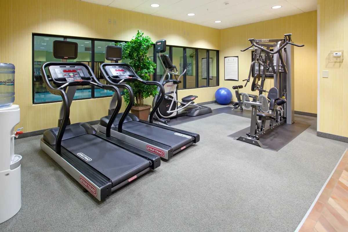 The fitness center is equipped with treadmills, an elliptical machine, a weight station, and a water cooler, all arranged in a bright, motivational environment. The space hints at both individual workouts and the possibility of group fitness activities.