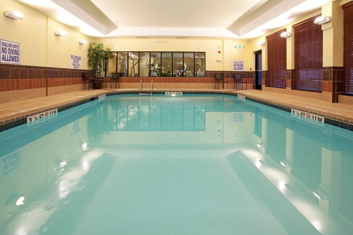 This interior image captures an inviting heated indoor swimming pool area. The pool is surrounded by comfortable seating, lush indoor plants, and exercise equipment visible in the background, creating a sense of relaxation and wellness. Guests are shown swimming, relaxing, and exercising.