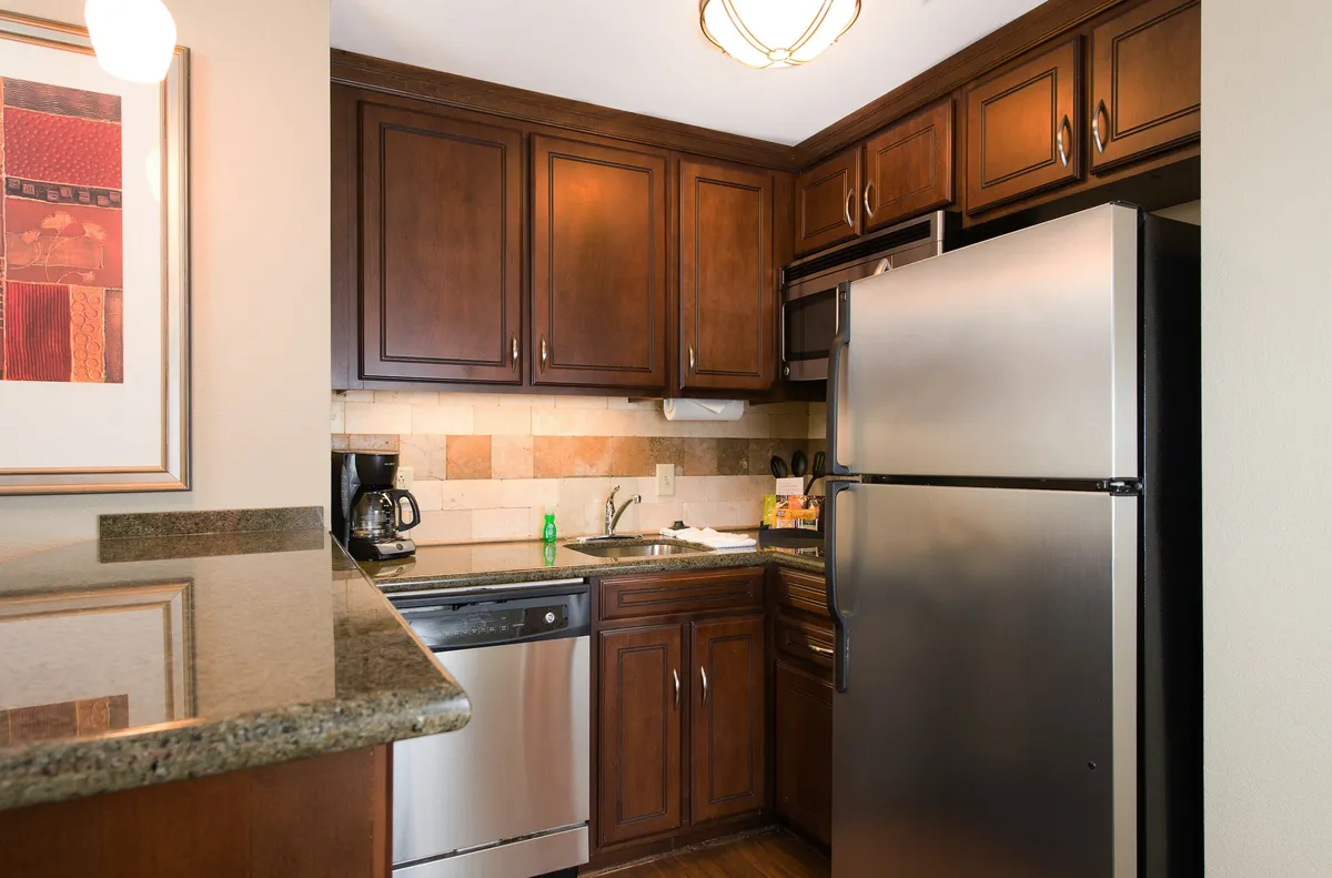 This warm, cozy interior shot features dark wooden cabinets, sleek stainless steel appliances, and granite countertops within a bright kitchen. The scene hints at both cooking and socializing, with brewing coffee and meal preparation activities subtly suggested.