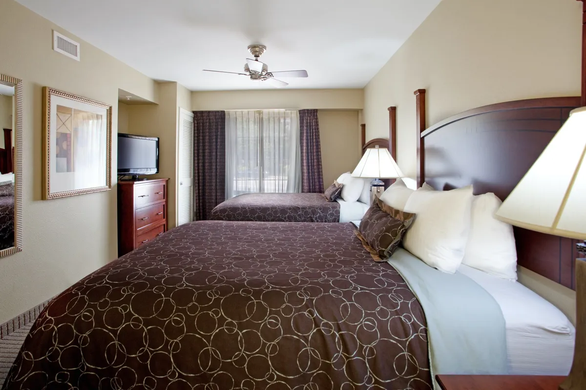 The second bedroom is shown with two cozy queen-sized beds, each topped with decorative pillows. Nightstands with lamps flank the beds, accompanied by a television and a dressing mirror, creating an inviting space for both relaxation and shared moments.