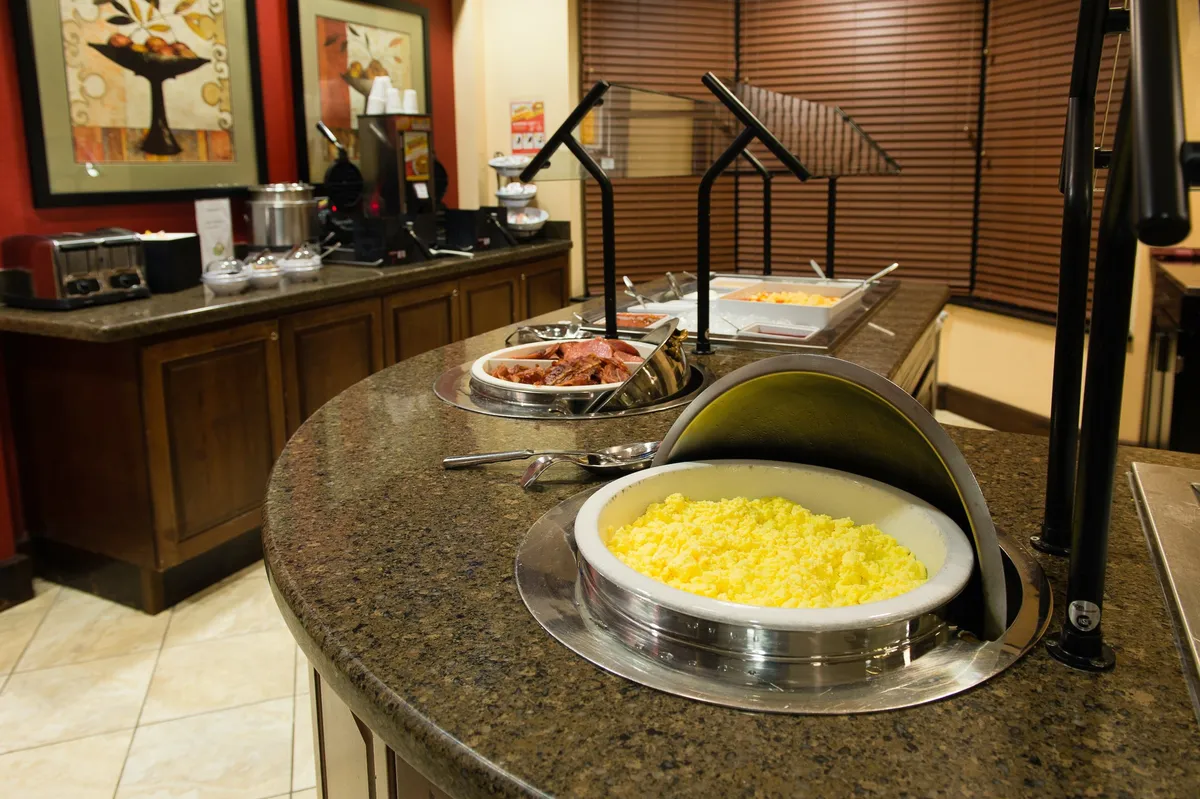 The image shows a welcoming breakfast buffet area set with a variety of food items on a long table. The space is decorated with contemporary artwork and features comfortable lighting, inviting guests to gather and enjoy a relaxing meal to start their day.